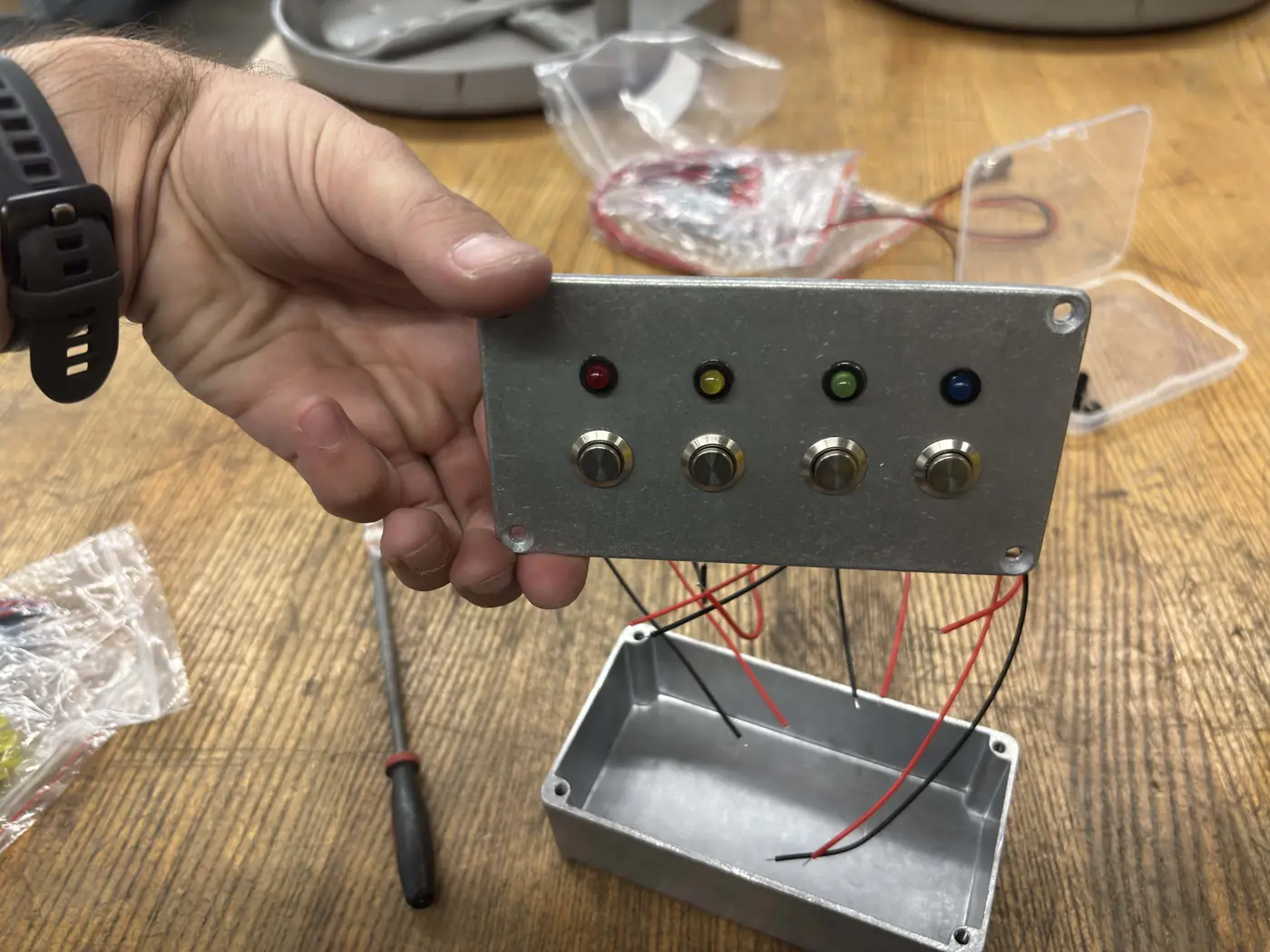 A hand holding a steel plate with four metal buttons and red, yellow, green, and blue LEDs, wired with red and black leads.