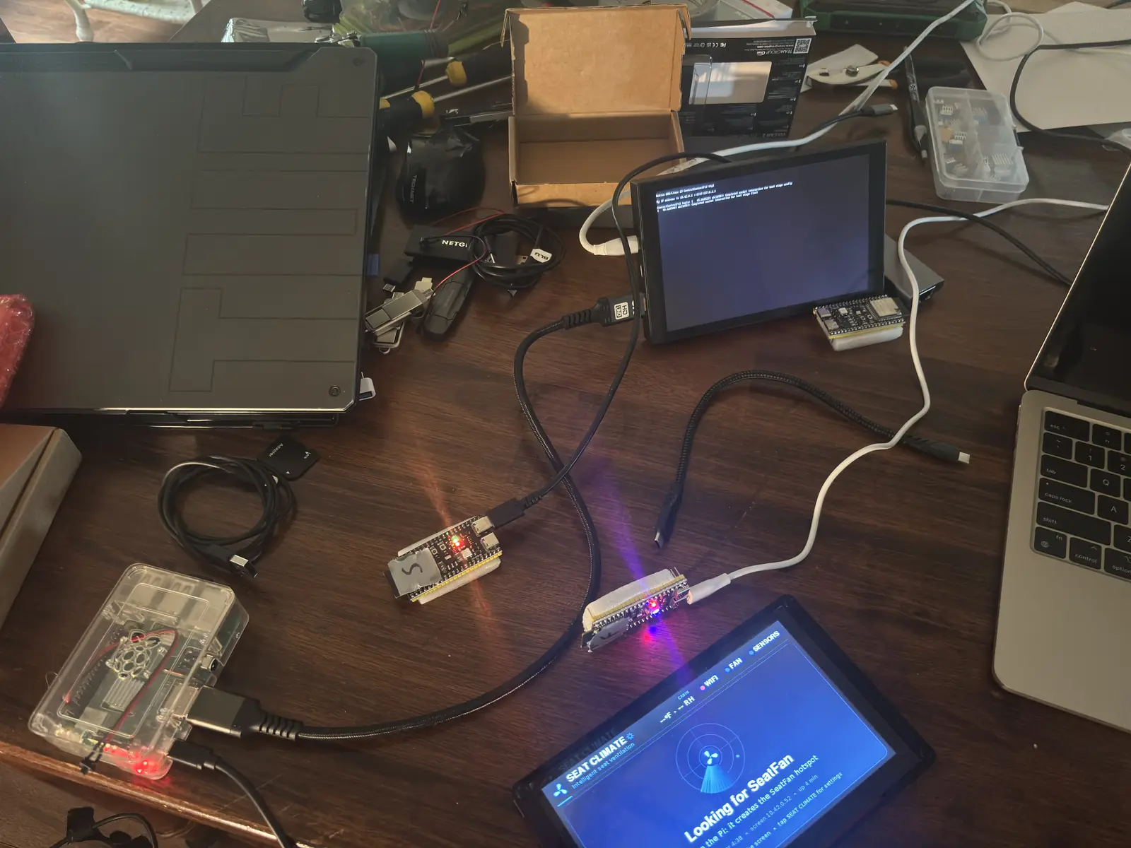 Workbench with a Raspberry Pi in a clear case, two ESP32 boards, and a touchscreen showing the Seat Climate page while it looks for the SeatFan hotspot.