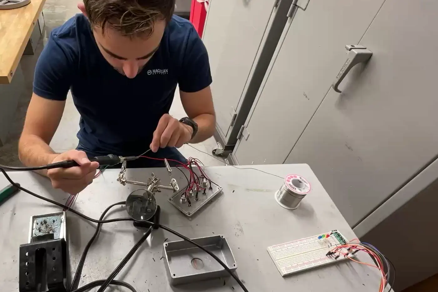 Eduardo Gomez soldering a lead held in a helping-hands clamp, with the button plate and a breadboard on the lab bench.