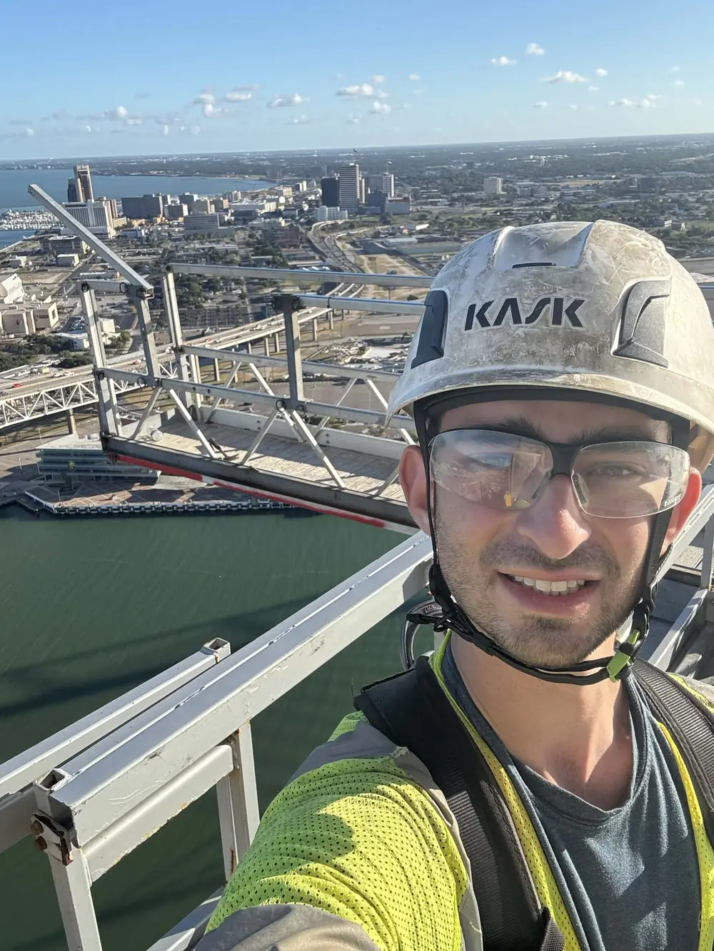 Eduardo Gomez on the Harbor Bridge tower in a hard hat and yellow vest, bay and skyline behind him.